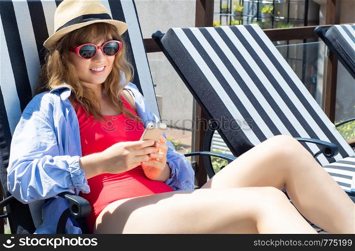 Young woman in hat using smartphone near the pool on beach chairs. Summer holidays