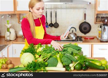 Young woman in kitchen having many green vegetables on table, holding tablet thinking about cooking something and searching for recipes in internet. Woman having green vegetables thinking about cooking