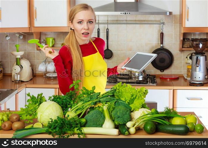 Young woman in kitchen having many green vegetables on table, holding tablet thinking about cooking something and searching for recipes in internet. Woman having green vegetables thinking about cooking