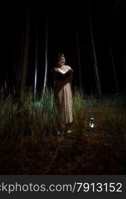 Young woman in nightgown reading book at night forest