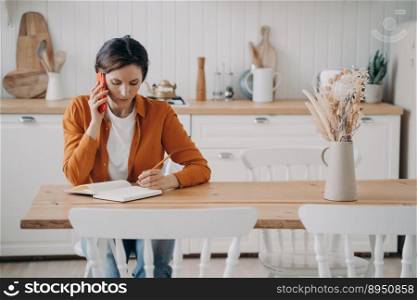 Young woman in orange shirt is freelancer. Hispanic girl is working from home sitting at her kitchen. Manager is receiving a phone call. Quarantine workplace and distance work concept.. Young woman is freelancer. Girl working from home and receiving a phone call. Quarantine workplace.