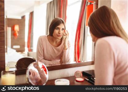 Young woman in pajama in front of the mirror in the bedroom. Female person cares for skin. Morning facial cleaning procedure