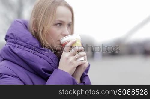 Young woman in purple jacket eating fast food and drinking hot tea outddor in cold weather.