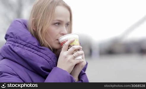 Young woman in purple jacket eating fast food and drinking hot tea outddor in cold weather.