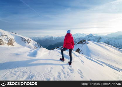 Young woman in red jacket in snowy mountains at sunset in winter. Landscape with beautiful girl on the hill in snow, rocks, blue sky at sunny day. Mountain pass in Dolomites, Italy. Tourism. Travel