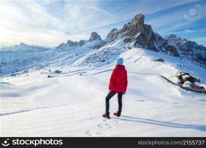 Young woman in red jacket in snowy mountains at sunset in winter. Landscape with beautiful girl on the hill in snow, rocks, blue sky at sunny day. Mountain pass in Dolomites, Italy. Tourism. Travel