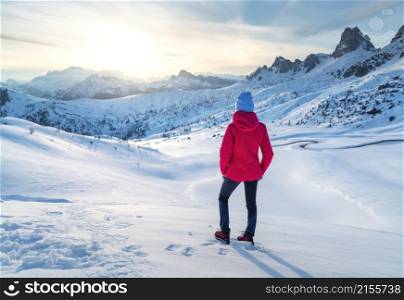 Young woman in red jacket in snowy mountains at sunset in winter. Landscape with beautiful girl on the hill in snow, rocks, blue sky at sunny day. Mountain pass in Dolomites, Italy. Tourism. Travel