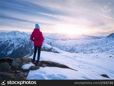 Young woman in red jacket in snowy mountains at sunset in winter. Landscape with beautiful girl on the hill in snow, rocks, colorful sky in evening. Mountain pass in Dolomites, Italy. Tourism. Travel