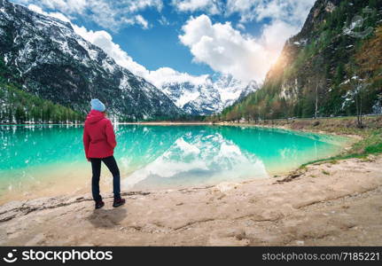 Young woman in red jacket is standing on shore of lake with azure water at sunny day in spring. Landscape with girl, reflection in water, snowy mountains, blue sky with clouds, green trees. Travel