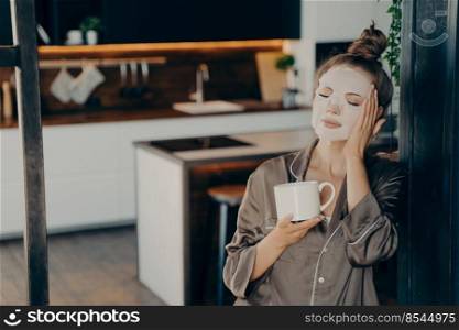Young woman in silk pajama staying at home and enjoying morning cup of coffee while applying white skincare disposable mask on face. Daily routine procedure for rejuvenation and care of skin. Young woman in silk pajamas staying at home and enjoying morning cup of coffee