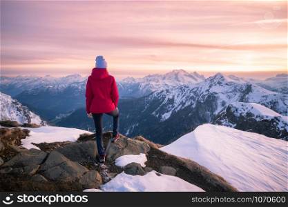 Young woman in snowy mountains at sunset in winter. Beautiful slim girl on the mountain peak against snow covered rocks and colorful red sky with clouds in the evening. Travel in Dolomites. Tourism
