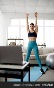 Young woman in sportswear at home, online fit training at the laptop. Female person doing exercise, internet sport workout, room interior on background. Young woman in sportswear, online fit training