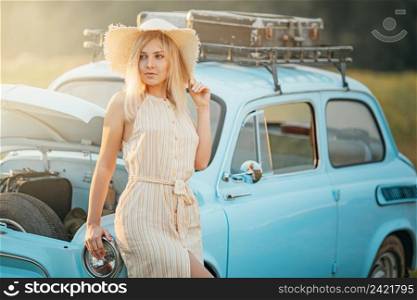 Young woman in straw hat standing near a blue vintage car with suitcases on a roof rack and opened hood, summer vibe