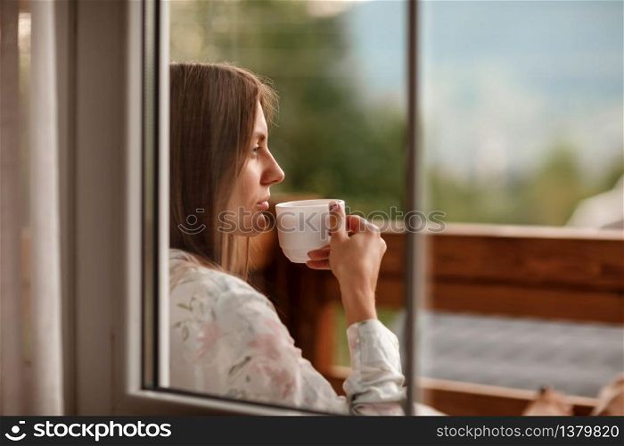 young woman in stylish nightwear enjoy drinking coffee or tea outdoor on balcony in the morning and looks at the mountains. young woman in stylish nightwear enjoy drinking coffee or tea outdoor on balcony in the morning and looks at the mountains.