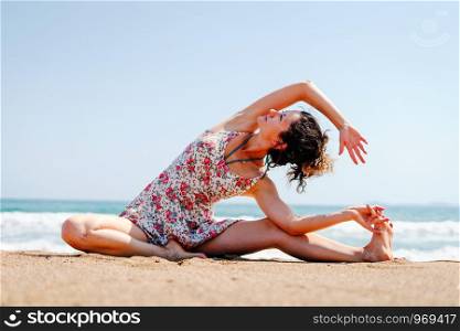 Young woman in summer dress practice yoga at the beach by the sea or ocean in sunny day stretching