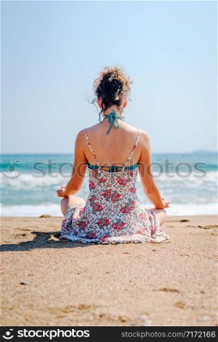Young woman in summer dress practice yoga meditating at the beach by the sea or ocean in sunny day stretching meditation relaxing meditate lotus position back view