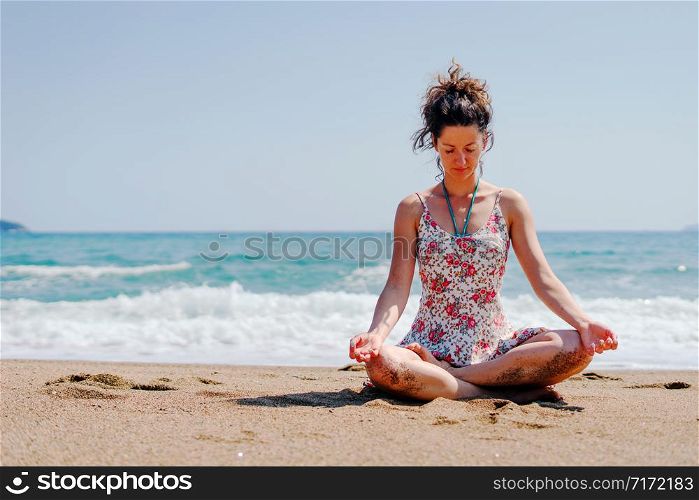 Young woman in summer dress practice yoga meditating at the beach by the sea or ocean in sunny day stretching meditation relaxing meditate lotus position