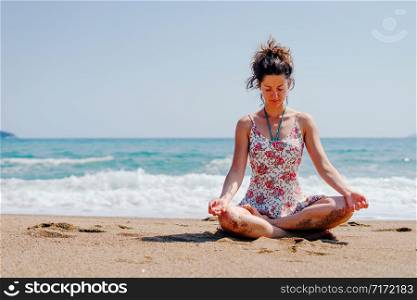 Young woman in summer dress practice yoga meditating at the beach by the sea or ocean in sunny day stretching meditation relaxing meditate lotus position