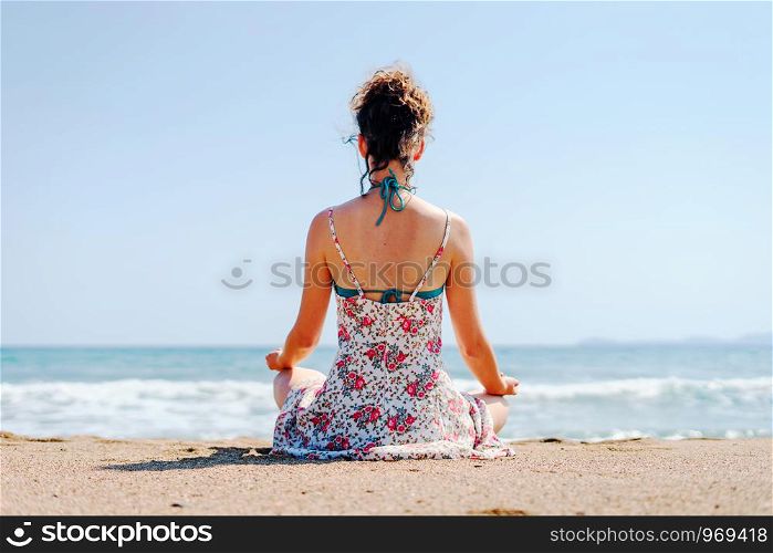 Young woman in summer dress practice yoga meditating at the beach by the sea or ocean in sunny day stretching meditation relaxing meditate lotus position back view