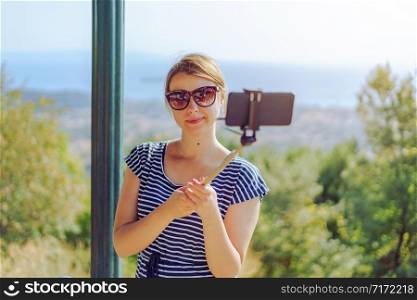 young woman in summer dress using selfie stick and her smart phone to take photo on the mountain above the sea while travel in september or october autumn or summer