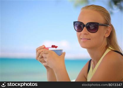 Young woman in sunglasses eating fruit ice-cream on the beach on a hot summer day. Bright blue sea and sky in background