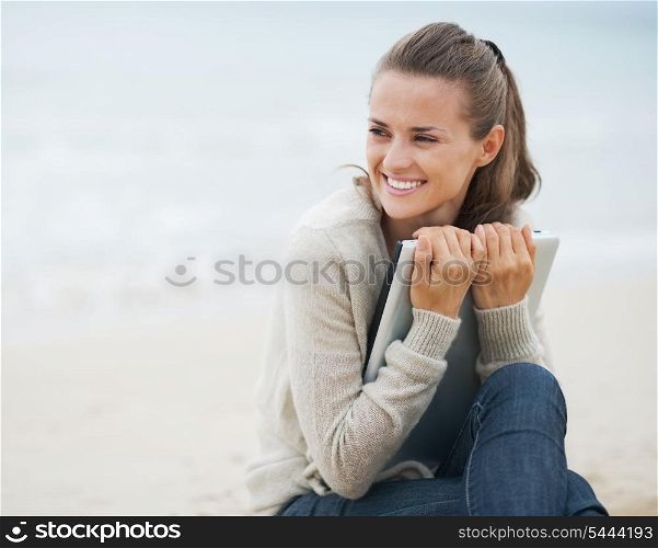 Young woman in sweater embracing laptop and looking on copy space while sitting on beach