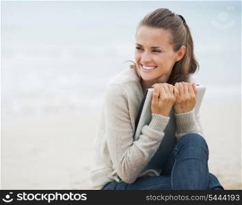Young woman in sweater embracing laptop and looking on copy space while sitting on beach