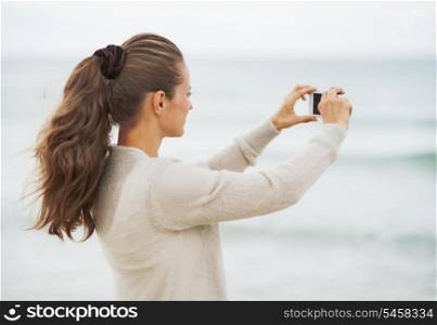 Young woman in sweater on beach taking photo using cell phone