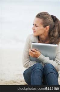 Young woman in sweater sitting on lonely beach and embracing laptop