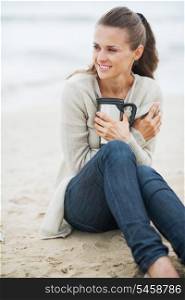 Young woman in sweater sitting on lonely beach with cup of hot beverage