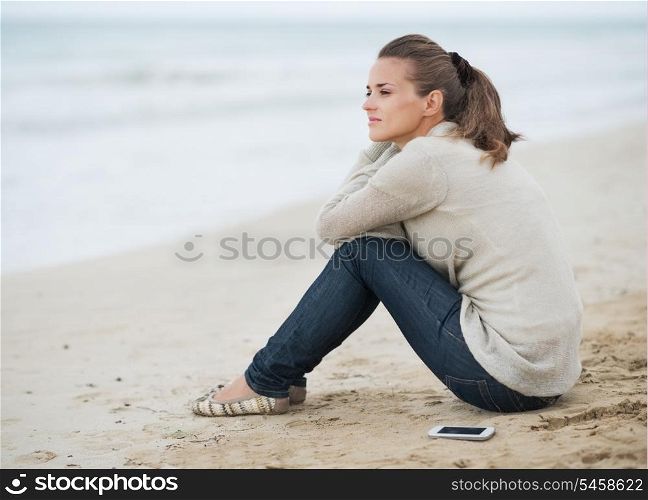 Young woman in sweater with cell phone sitting on lonely beach and looking into distance