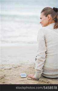 Young woman in sweater with cell phone sitting on lonely beach . rear view