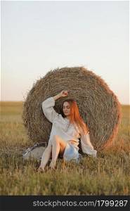 young woman in the beautiful light of the summer sunset in a field is sitting near the straw bales. beautiful romantic girl with long hair outdoors in field.. young woman in the beautiful light of the summer sunset in a field is sitting near the straw bales. beautiful romantic girl with long hair outdoors in field