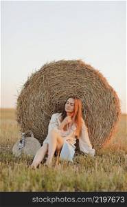 young woman in the beautiful light of the summer sunset in a field is sitting near the straw bales. beautiful romantic girl with long hair outdoors in field.. young woman in the beautiful light of the summer sunset in a field is sitting near the straw bales. beautiful romantic girl with long hair outdoors in field