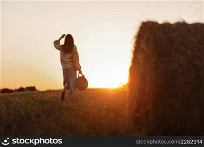 young woman in the beautiful light of the summer sunset in a field is walking near the straw bales. beautiful romantic girl with long hair outdoors in field. young woman in the beautiful light of the summer sunset in a field is walking near the straw bales. beautiful romantic girl with long hair outdoors in field.