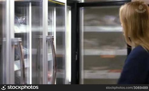 Young woman in the frozen section opening the fridge, choosing and taking the product