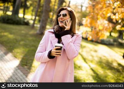 Young woman in the pink coat talking over the smartphone and holding coffee to go in hand at autumn park