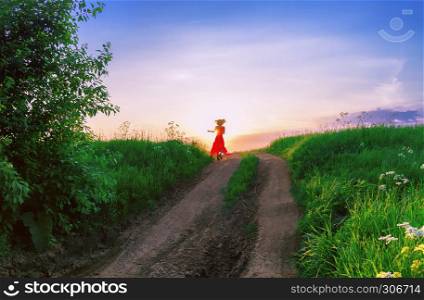 Young woman in the red dress is dancing far on a dirt path in a green field in the rays of the setting sun.. Young Woman Dancing Against The Setting Sun