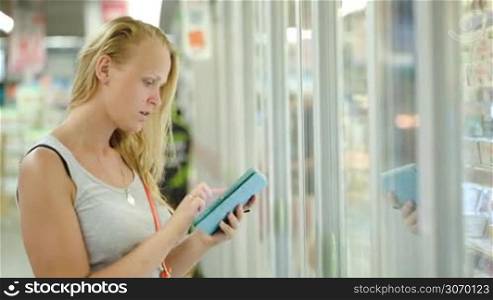 Young woman in the store checking list of products on touch pad and taking some from fridge