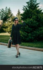 young woman in vintage black polka dot dress with retro suitcase in hand posing outside
