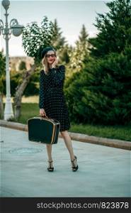 young woman in vintage black polka dot dress with retro suitcase in hand posing outside