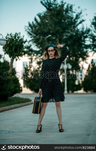 young woman in vintage black polka dot dress with retro suitcase in hand posing outside