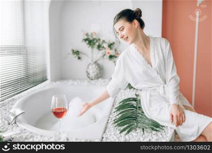 Young woman in white bathrobe sitting on the edge of the bath with foam. Bathroom interior with window and glass with red wine on background