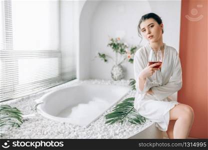 Young woman in white bathrobe sitting on the edge of the bath with foam. Bathroom interior with window and glass with red wine on background. Young woman in n white bathrobe, bathroom interior