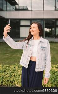 Young woman in white denim jacket and crop top taking selfie on street, office building in the background. Streetwear casual clothes, using technology concept. Young woman in white denim jacket taking selfie on street. Office building in the background