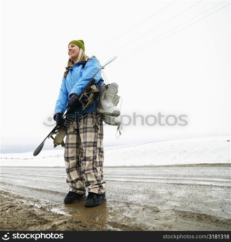 Young woman in winter clothes standing in muddy dirt road holding snowboard and boots.