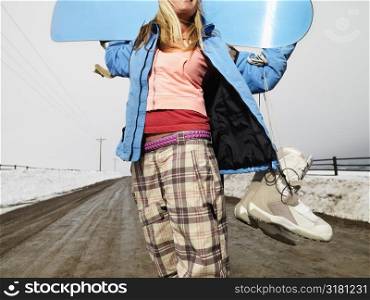 Young woman in winter clothes standing on muddy dirt road holding snowboard and boots.