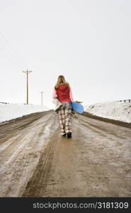 Young woman in winter clothes walking alone down muddy dirt road carrying snowboard and boots.