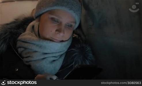 Young woman in winter overclothes browsing web on cell when traveling by bus in night city