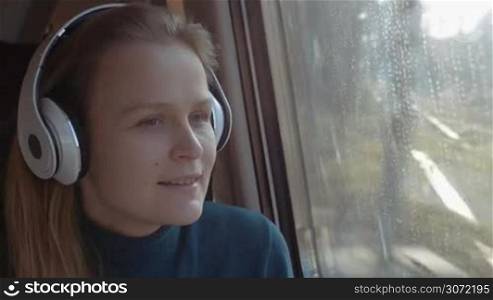 Young woman in wireless earphones relaxing with music in the train. She singing song, dancing and enjoying scene from the window